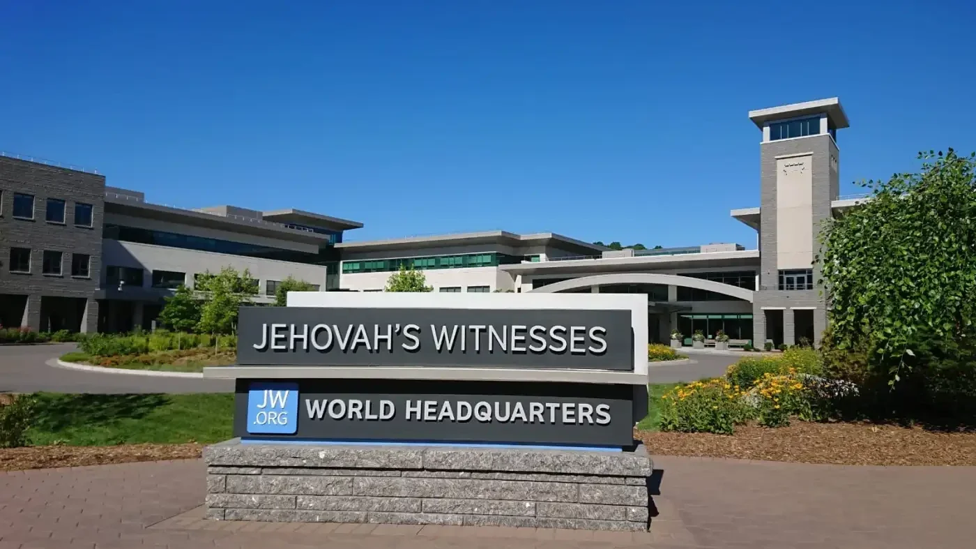 Cartel de la sede mundial de los Testigos de Jehová en Warwick, Nueva York, con los edificios al fondo.