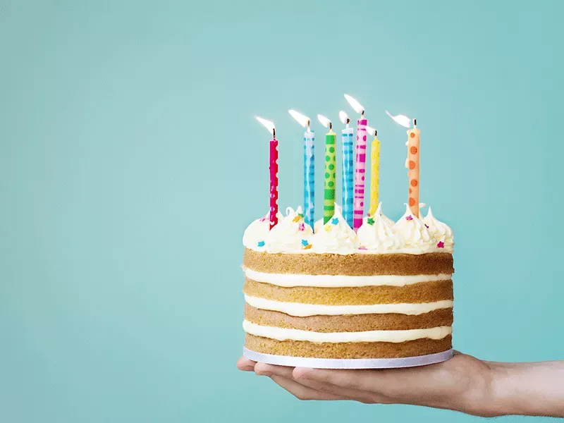 Una mano sostiene una tarta de cumpleaños con velas de colores encendidas sobre un fondo turquesa.