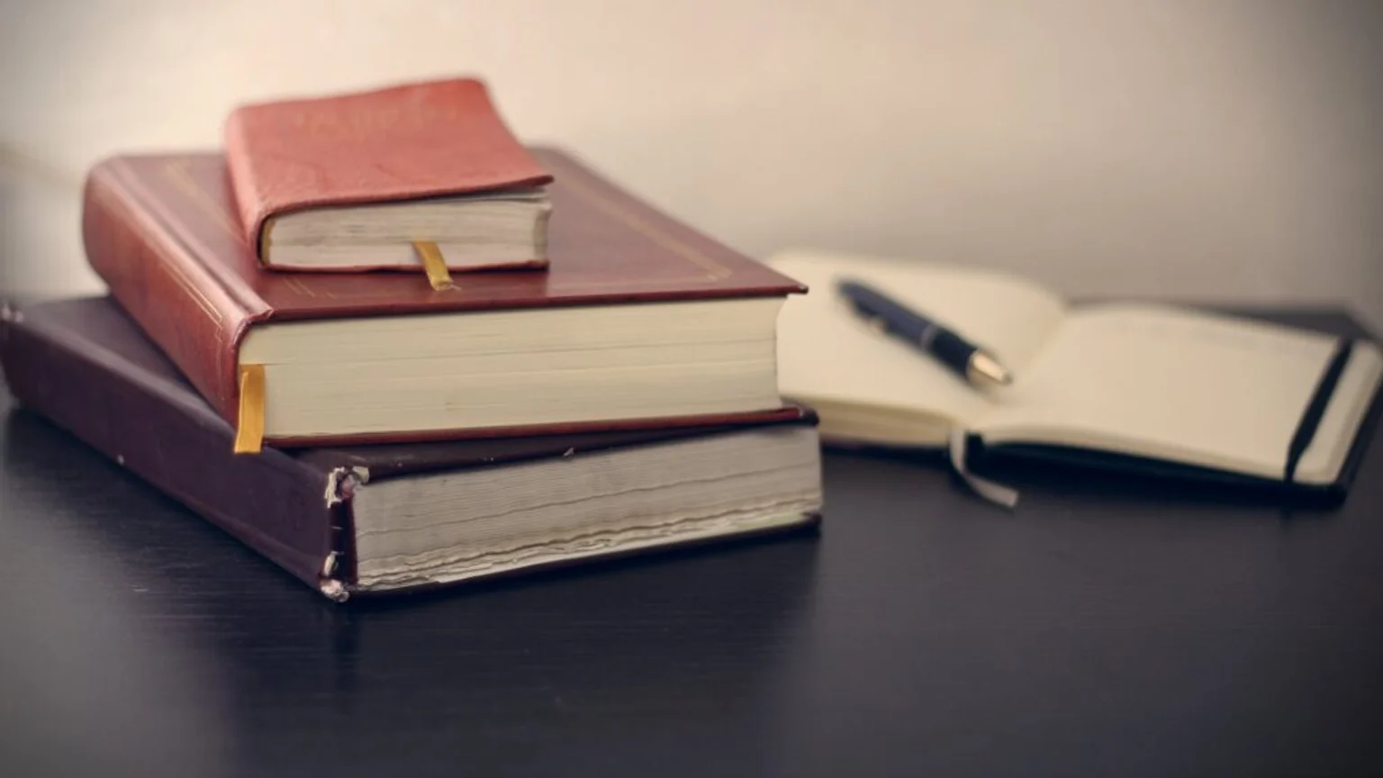 Libros de tapa antigua apilados junto a un cuaderno abierto y un bolígrafo.